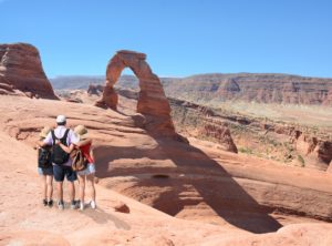 People hiking on vacation. Father standing with arms around his family on top of the mountain, looking at beautiful summer mountains landscape. Blue sky in the background. Arches National Park, Moab, Utah.